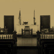 Empty courtroom with wooden benches, a judge’s bench, jury seats, and American and state flags in the background.