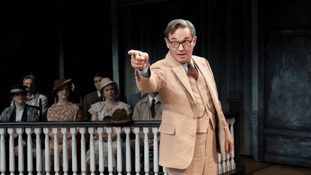 A man in a beige suit stands in front of a seated group, pointing forward, with a wooden railing and dark background behind him, suggesting a courtroom or theatrical scene.