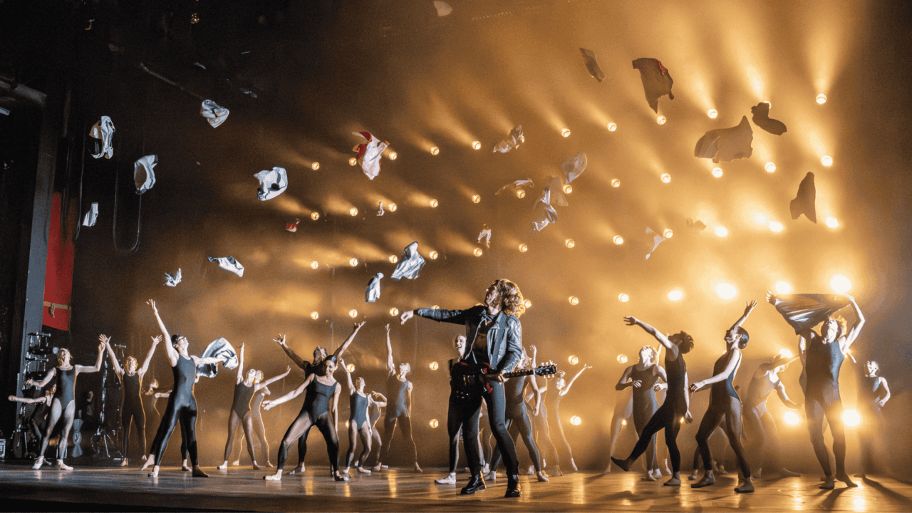 A group of dancers perform energetically on stage as clothes are thrown into the air, with dramatic lighting and a guitarist in the center.