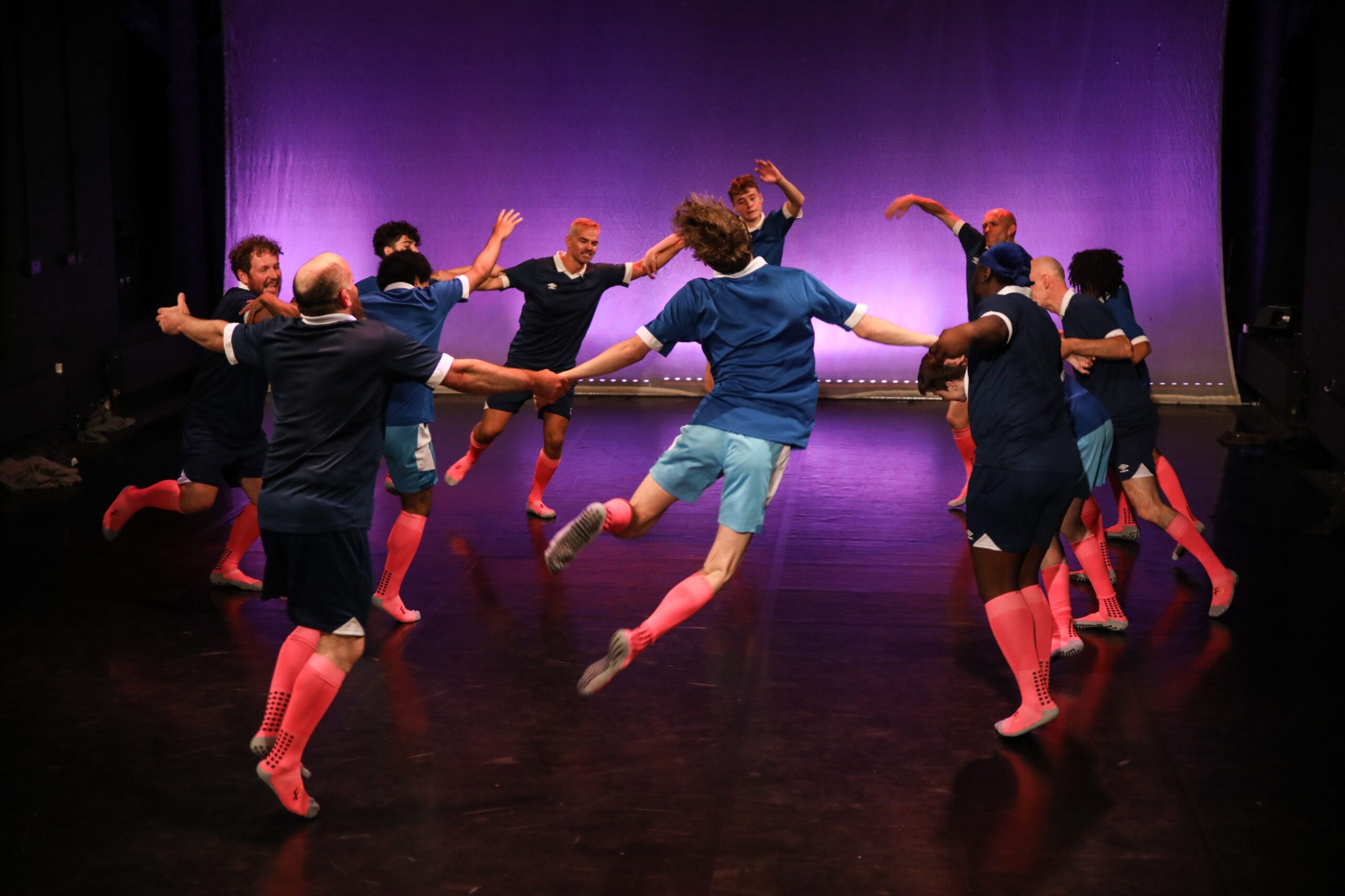 A group of people in blue shirts and pink knee socks hold hands in a circle, jumping in unison on a stage with a purple-lit backdrop.