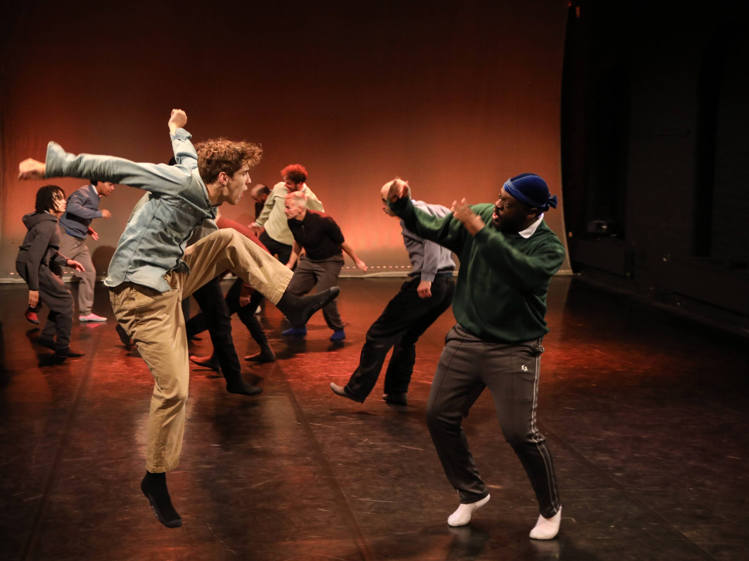 A group of people perform a contemporary dance on stage, with one person jumping and raising a knee while another faces them with raised arms.
