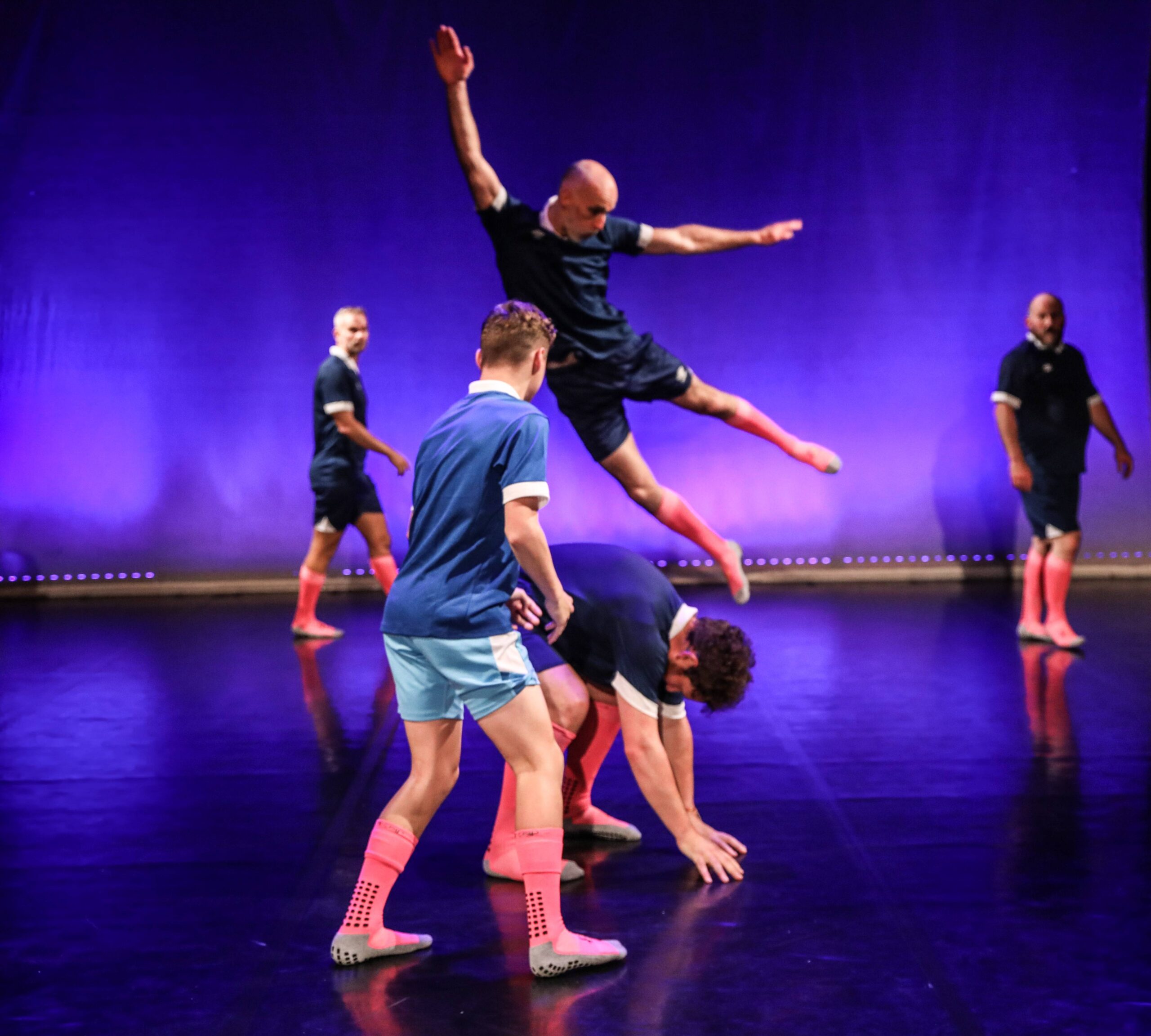 Five people in athletic outfits perform a dance routine on stage; one person leaps over another who is bent down, with others watching. The background is lit with purple lighting.