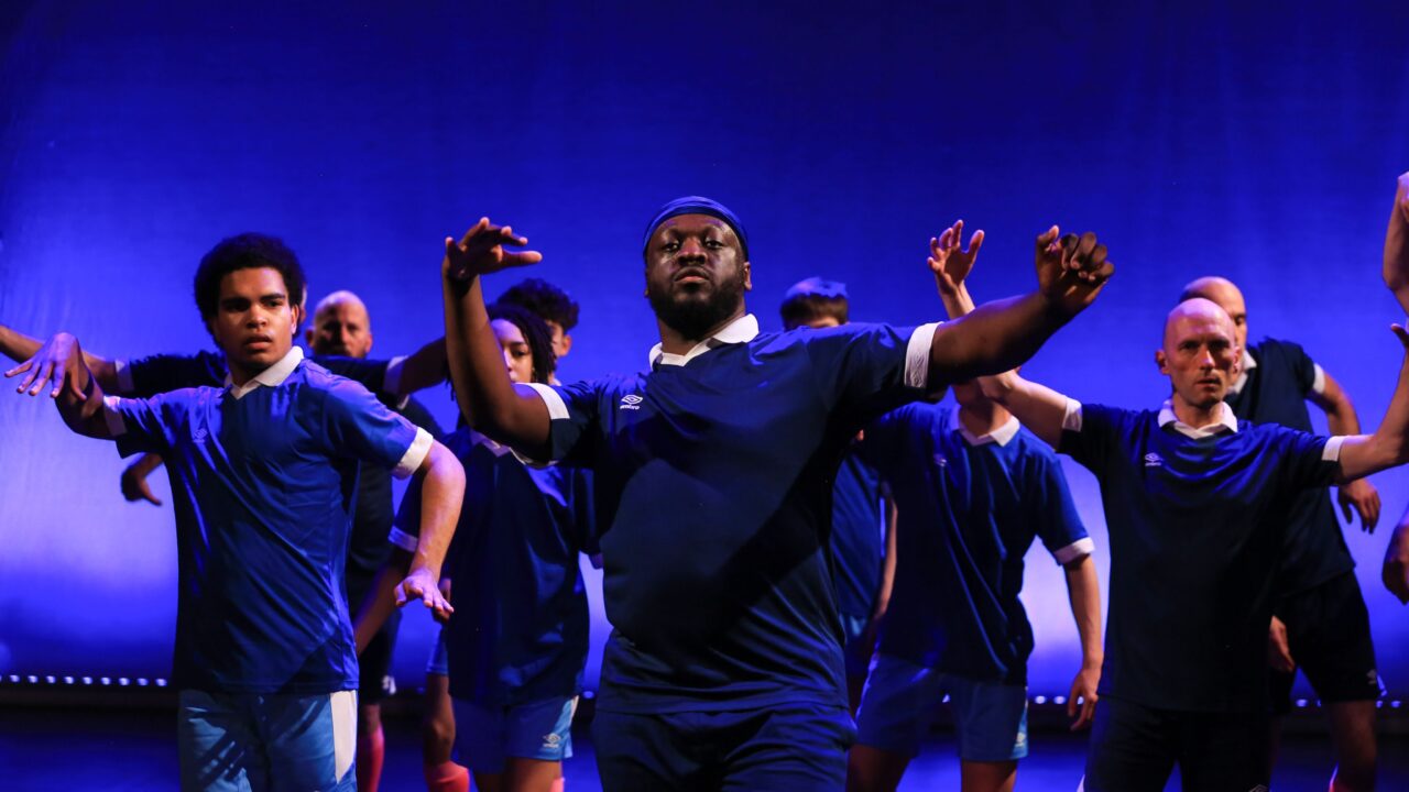 A group of people in blue athletic shirts performs a choreographed dance on stage with blue lighting in the background.