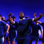 A group of people in blue athletic shirts performs a choreographed dance on stage with blue lighting in the background.