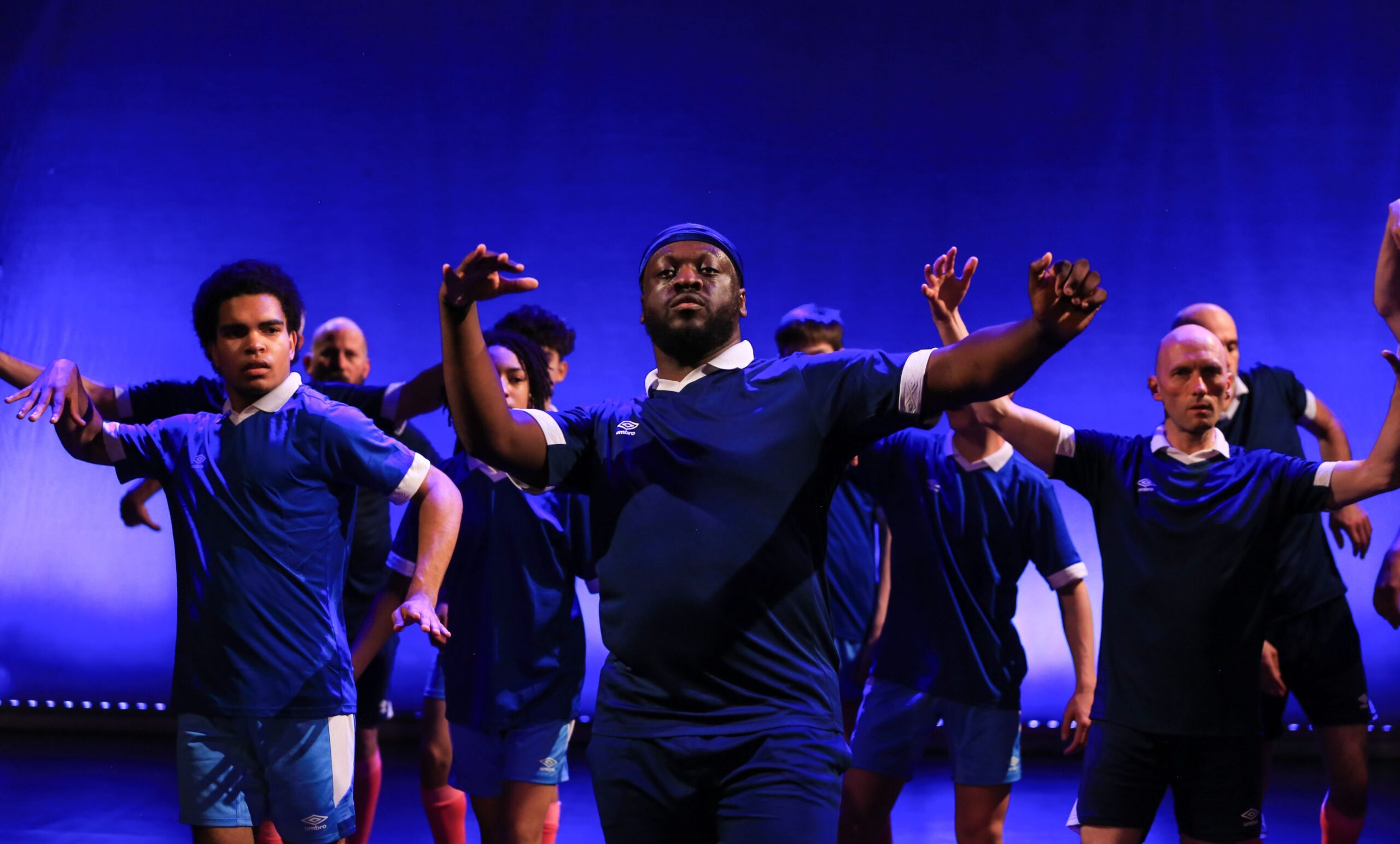 A group of people in blue athletic shirts performs a choreographed dance on stage with blue lighting in the background.