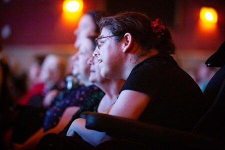 A group of people seated in a dimly lit theater, watching a performance, with attentive expressions.