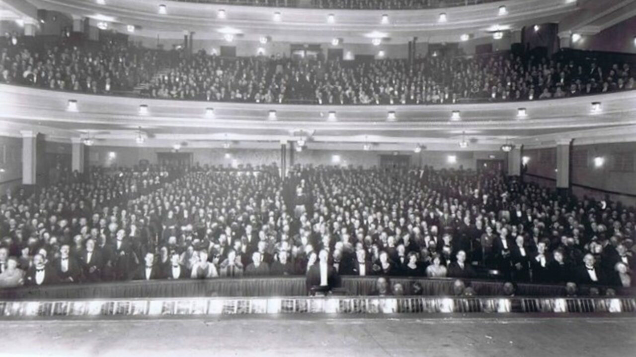 A large audience sits in a packed, multi-tiered theatre, facing the stage, with all seats occupied and people dressed formally.