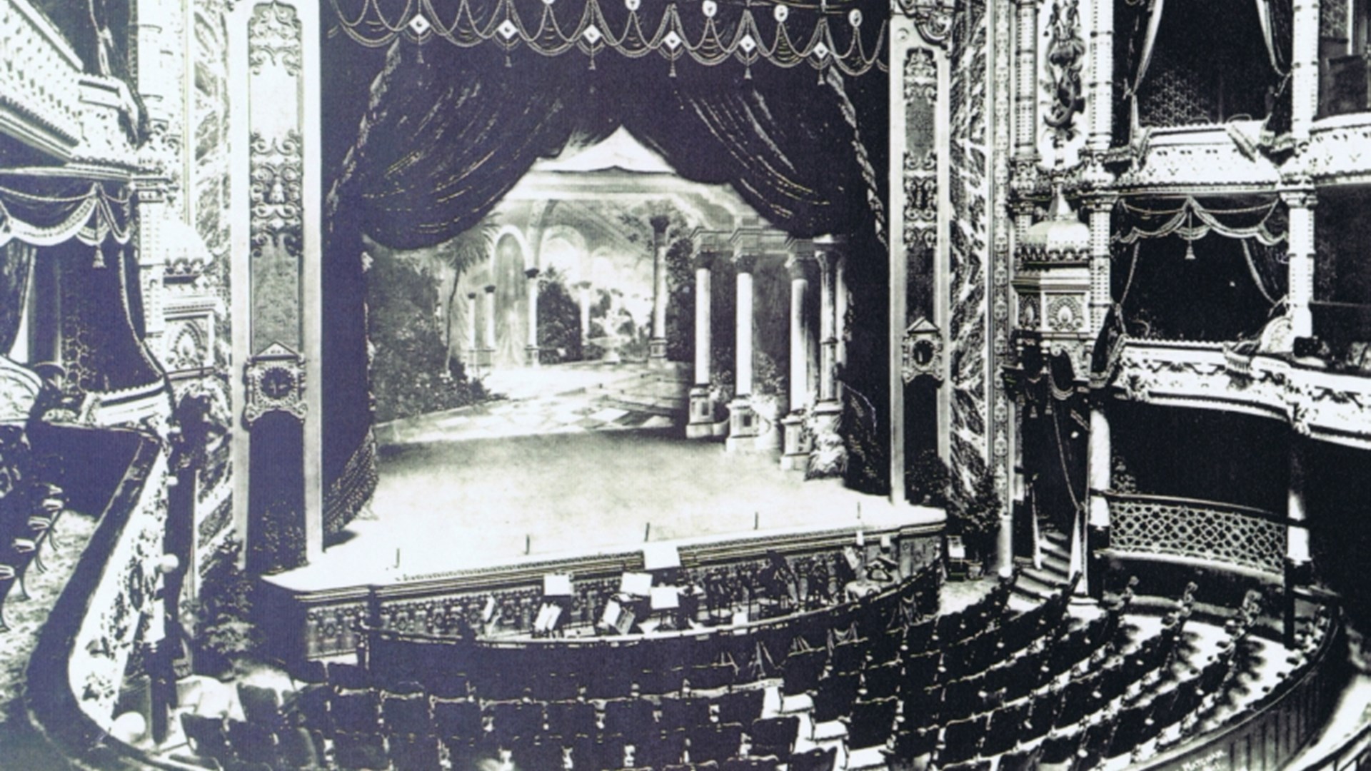 Black and white photo of an ornate theatre with decorative balconies, empty seats, and a stage set with classical columns and a scenic backdrop.