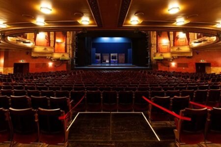 Empty theater auditorium with red seats facing a stage set with blue scenery, surrounded by ornate decor and overhead lighting.