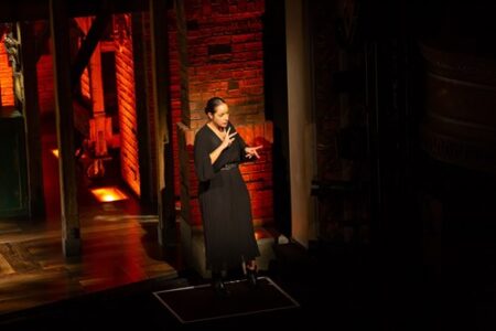 A person in a black dress interprets a theatre performance with BSL against a backdrop of red brick walls, with warm lighting.