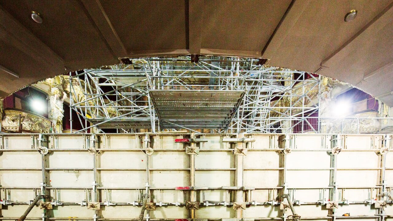A large metal scaffolding structure is set up inside a building with ornate details, partially blocked by a temporary construction barrier.
