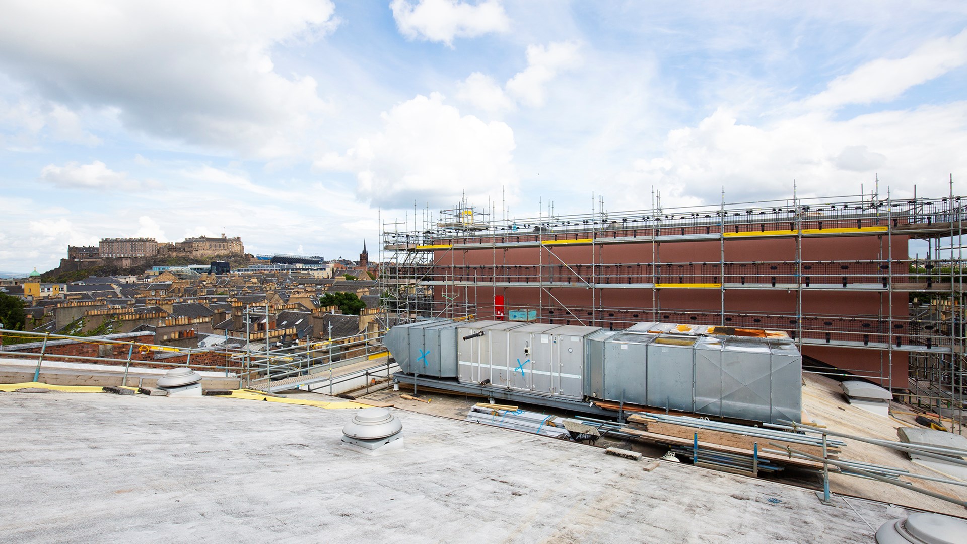 View of a rooftop construction site with scaffolding and ventilation units, overlooking a cityscape with a historic castle in the distance under a partly cloudy sky.