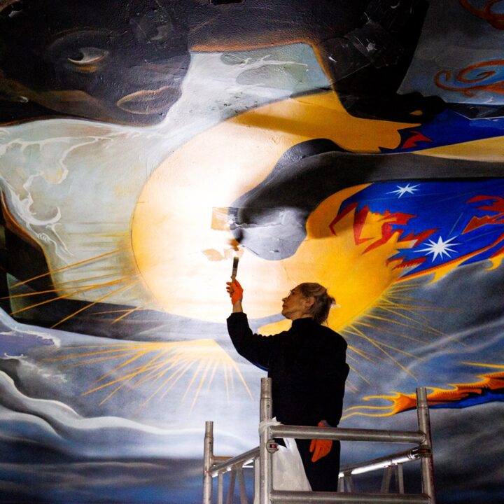 A person stands on scaffolding, restoring a large, colourful mural on a ceiling with a paintbrush.