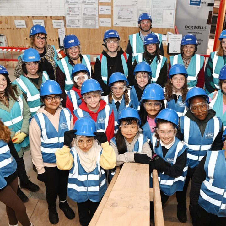 A group of children and adults wearing blue hard hats and reflective vests pose together inside a construction site.