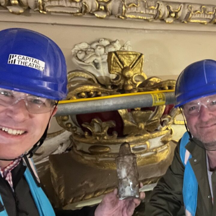 Two men in blue hard hats and safety vests pose in front of ornate gold architectural details; one holds a small glass bottle.