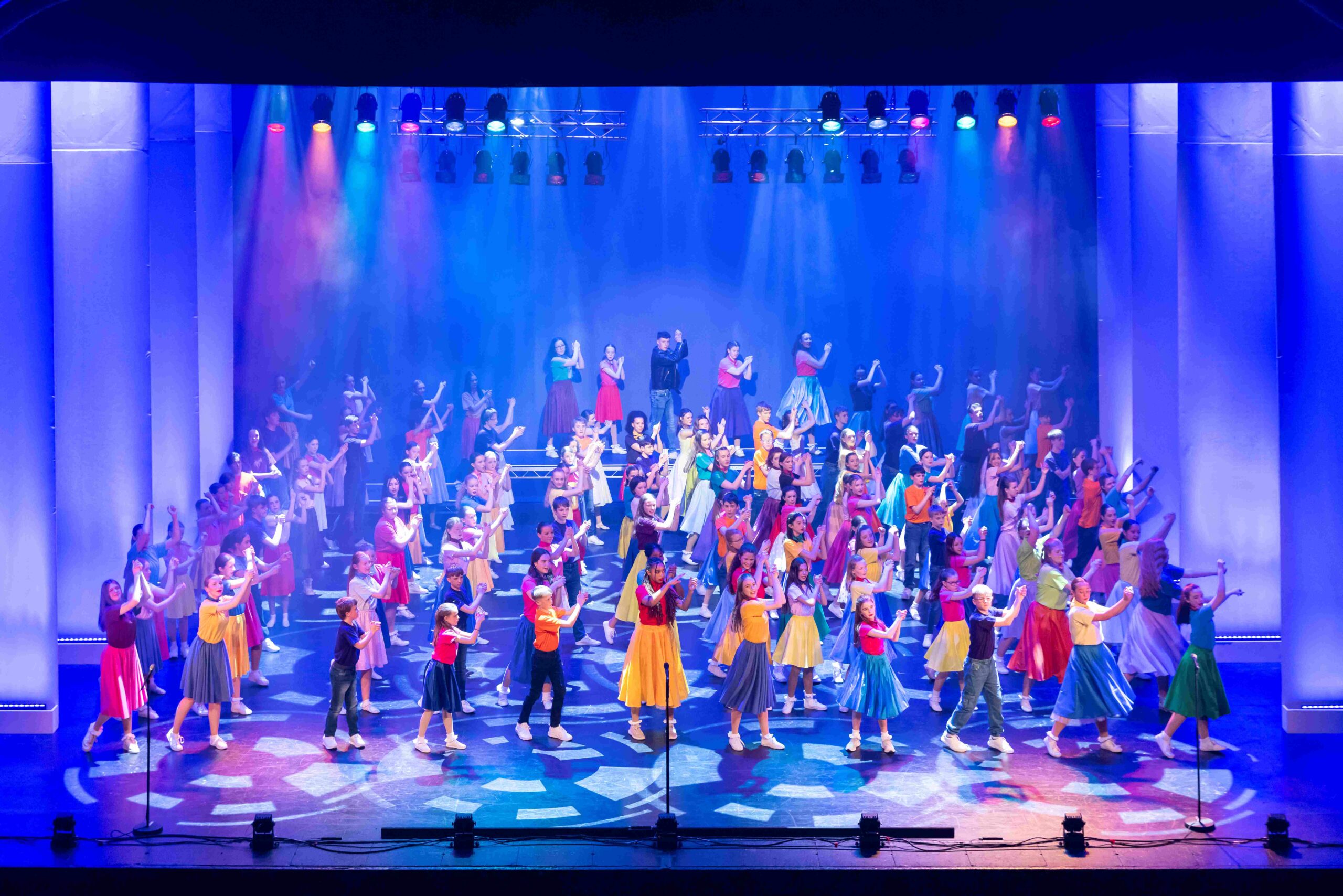Large group of performers in colorful costumes dance and gesture on a brightly lit stage under multicolored lights.