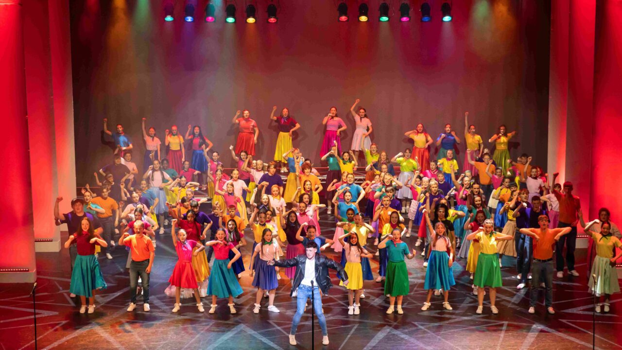 A large group of people in colorful costumes perform a choreographed dance on a brightly lit stage with red and purple lighting.