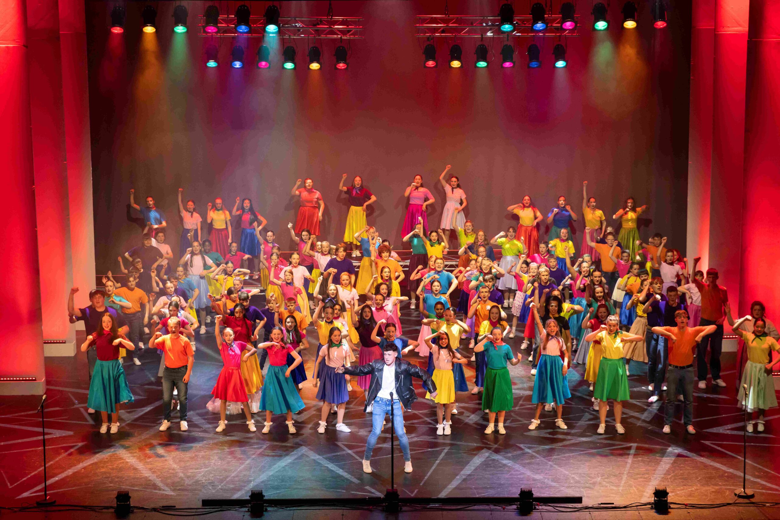 A large group of people in colorful costumes perform a choreographed dance on a brightly lit stage with red and purple lighting.