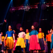 A group of performers on stage, with one person in a leather jacket singing in front and others in colorful skirts and tops standing behind.