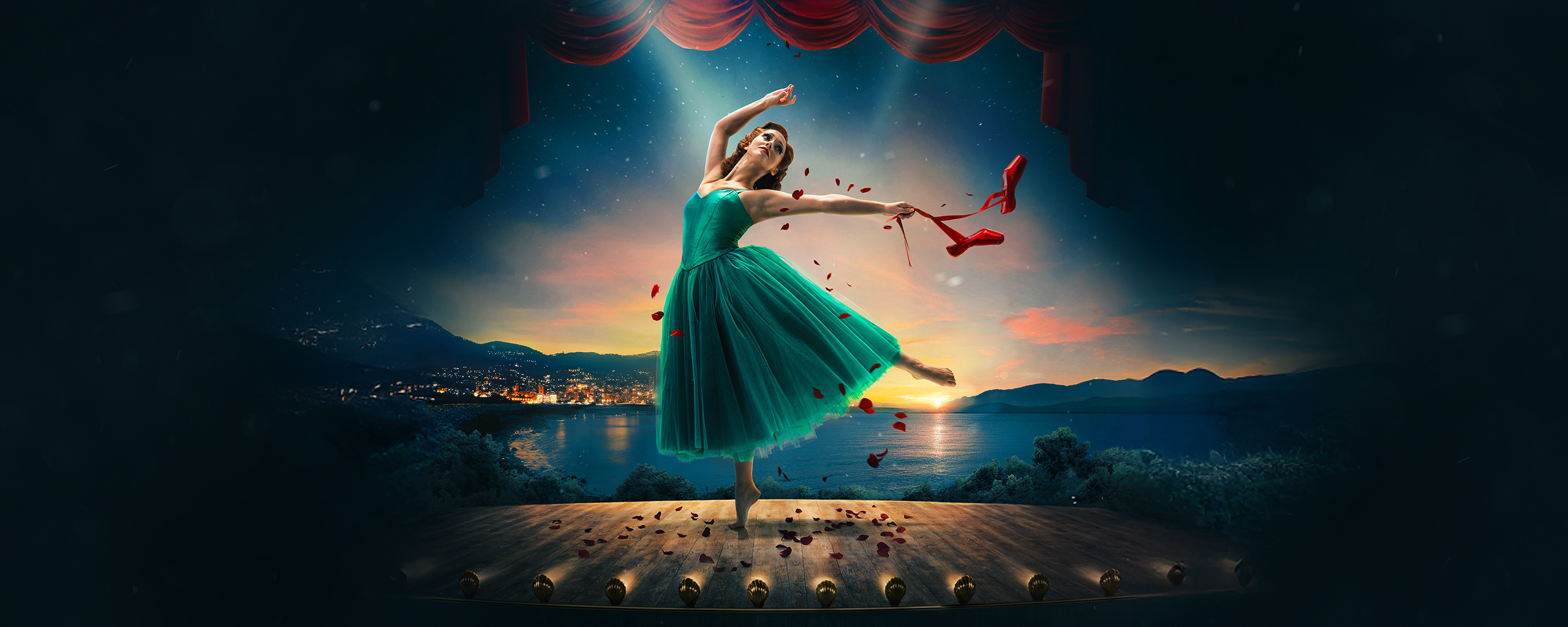 A ballerina in a green dress dances on stage at sunset, holding red ballet shoes and surrounded by red rose petals, with a lakeside town and mountains in the background.