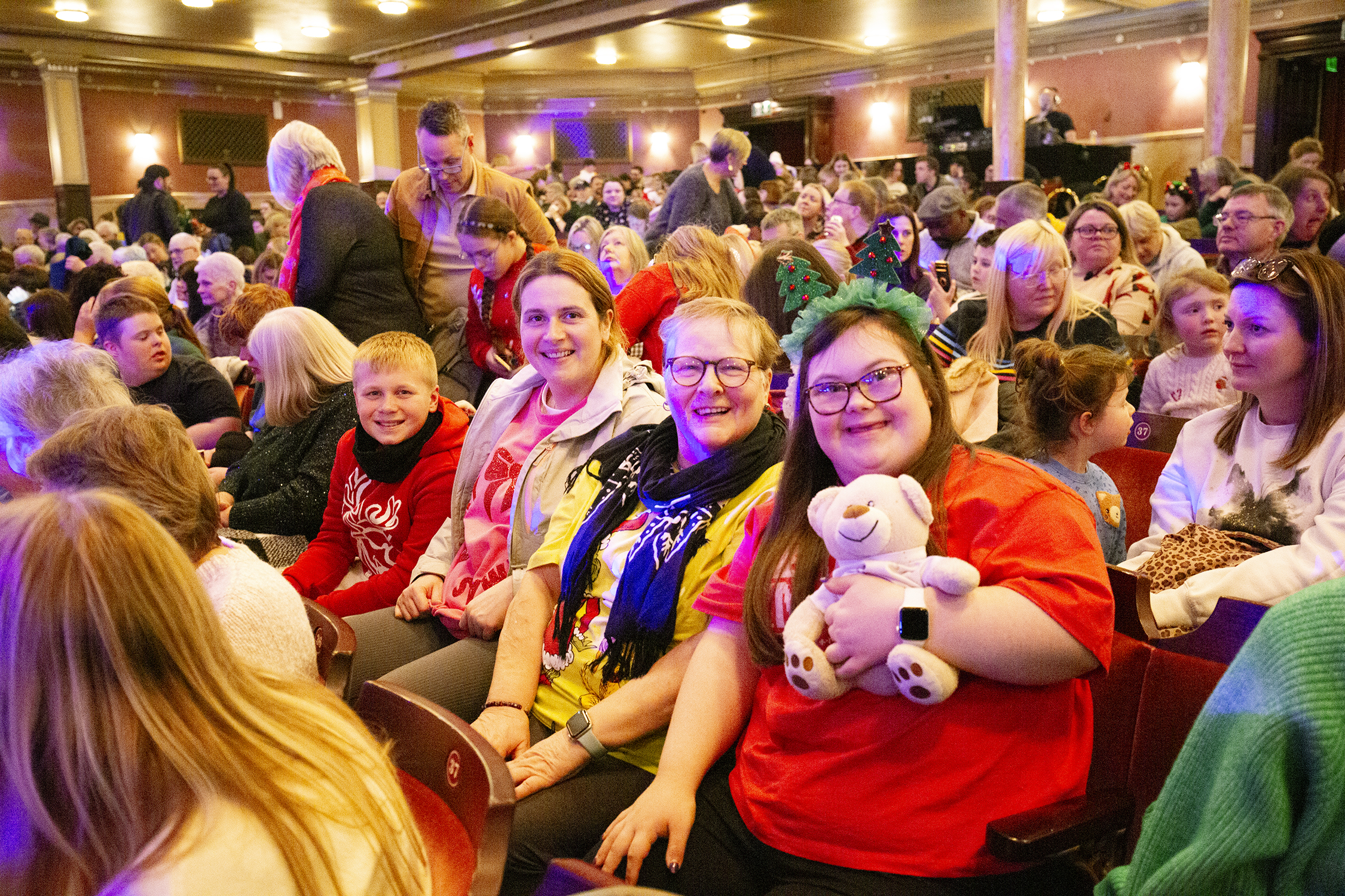 Audience seated in a theatre, waiting for a show to start. A group in the foreground smiles at the camera; one person holds a teddy bear and wears decorative headbands.