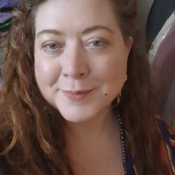 A woman with long wavy brown hair and fair skin is smiling at the camera. She is wearing a colorful top and gold earrings.