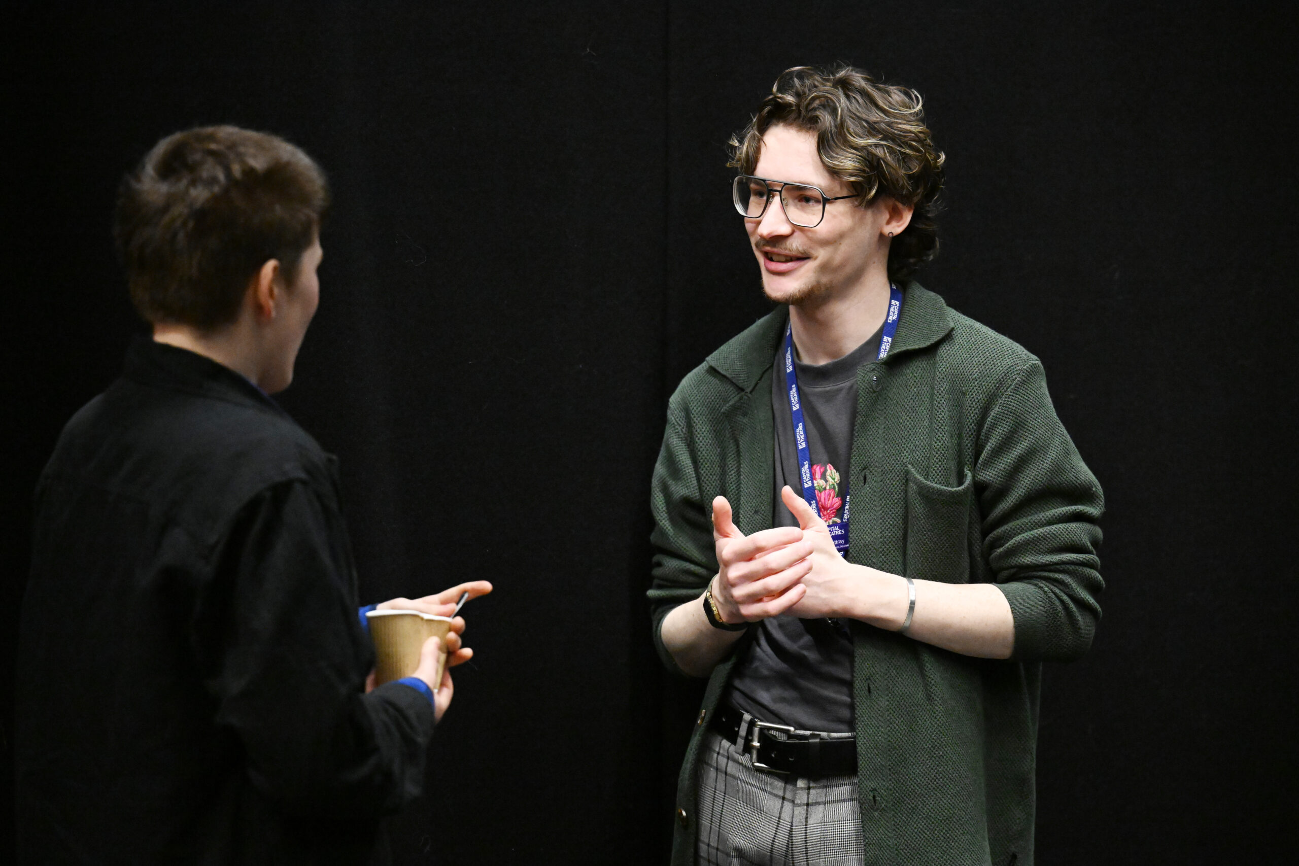 Two people engaged in conversation, one holding a cup. The person facing the camera gestures with their hands, wearing glasses and a lanyard, against a plain black background.