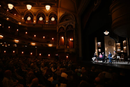 A panel of five people sits on a stage in a dimly lit theater, addressing an audience.