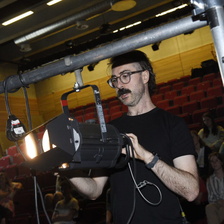 A man adjusts a stage spotlight in a theater with red seats, while a few people observe in the background.