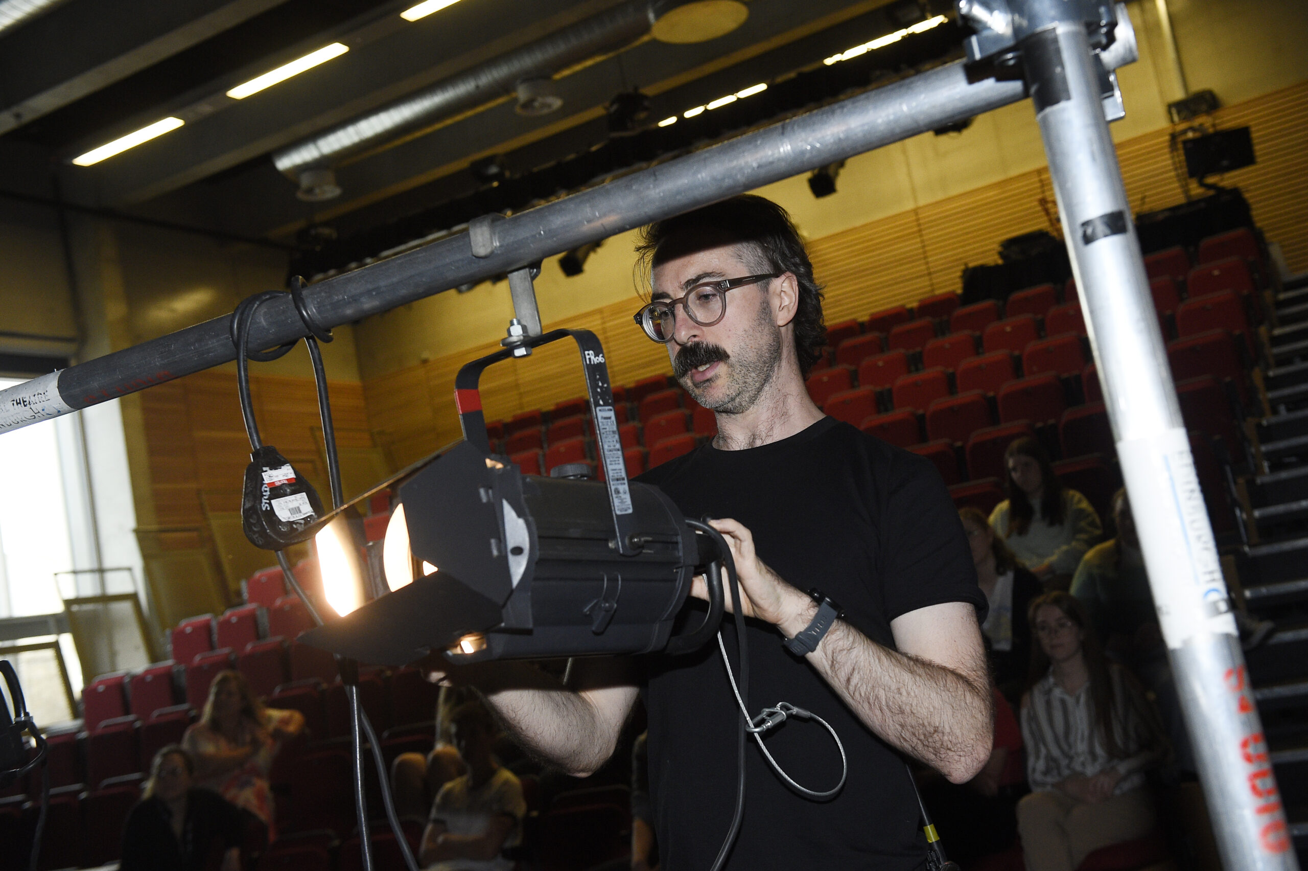 A man adjusts a stage spotlight in a theater with red seats, while a few people observe in the background.