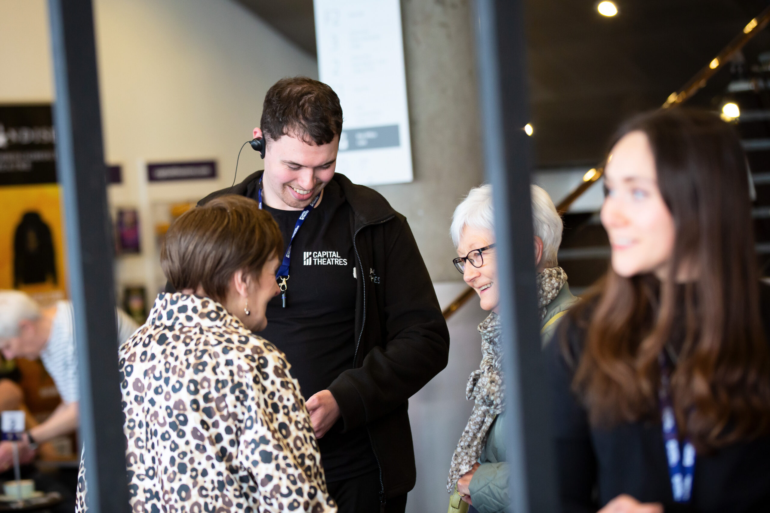 An usher wearing a headset and staff badge smiles while speaking with three women in the theatre foyer.