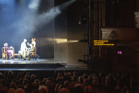 Image showing a dimly lit stage with a captioning screen on the right. The audience is in the foreground.