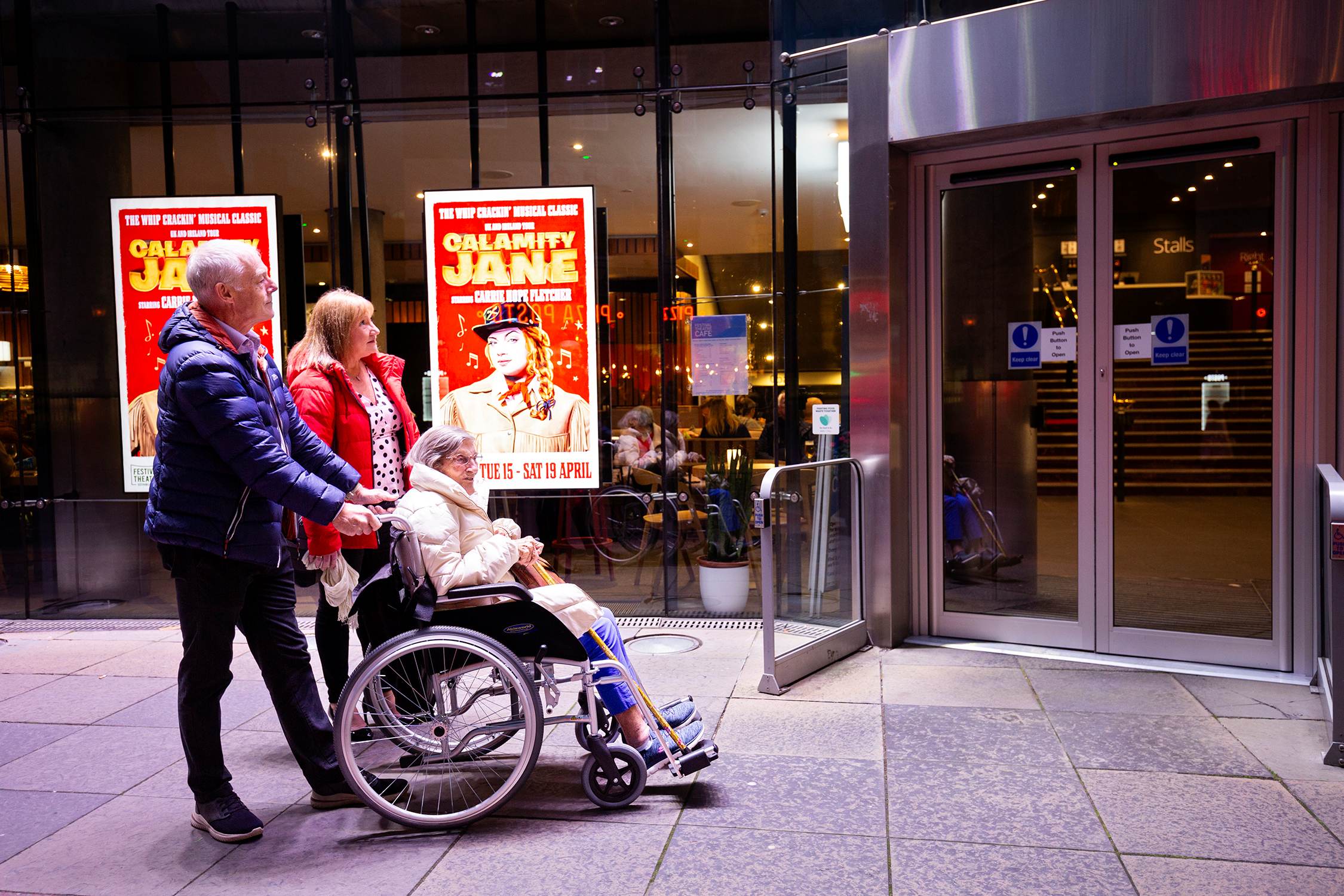 Two people are pushing a person in a wheelchair outside a building with glass doors. A poster for "Calamity Jane" is displayed in the background.