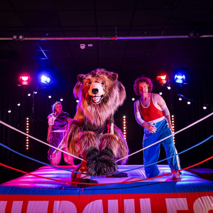A large bear costume character and two performers stand in a brightly lit wrestling ring with “Hercules” on the apron, surrounded by stage lights.