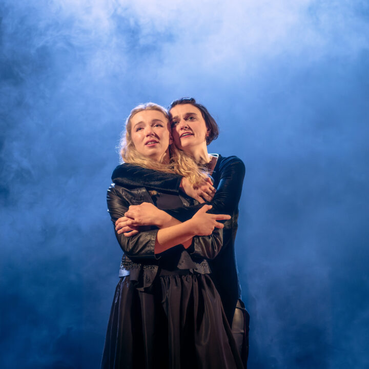 Two people stand closely together, with one person embracing the other from behind, both looking ahead with serious expressions against a smoky blue background.