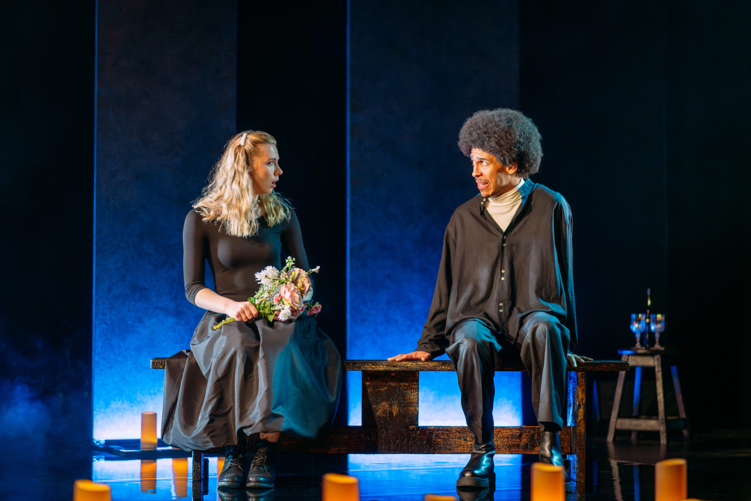 Two people sit on a bench onstage; the woman holds a bouquet of flowers, while the man looks at her. The lighting is dramatic, with candles on the floor and blue light in the background.
