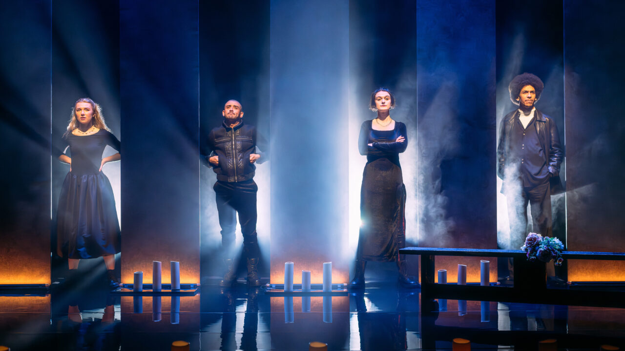 Four people stand on a dimly lit stage with dramatic lighting, surrounded by vertical light columns and candles, facing forward with serious expressions.