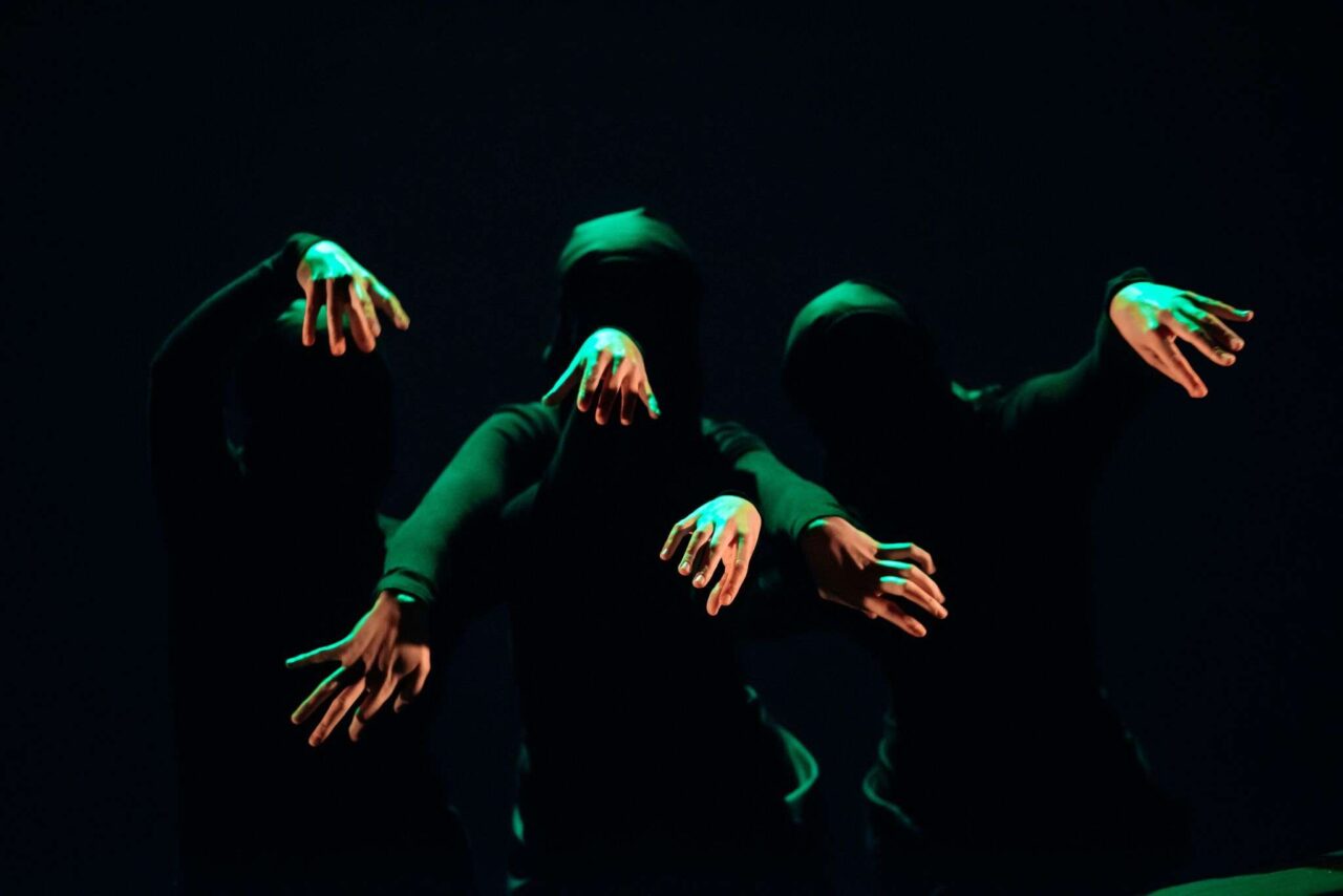 Three people in black clothing and masks extend their arms forward in dim green light against a dark background.