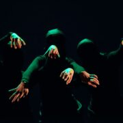 Three people in black clothing and masks extend their arms forward in dim green light against a dark background.