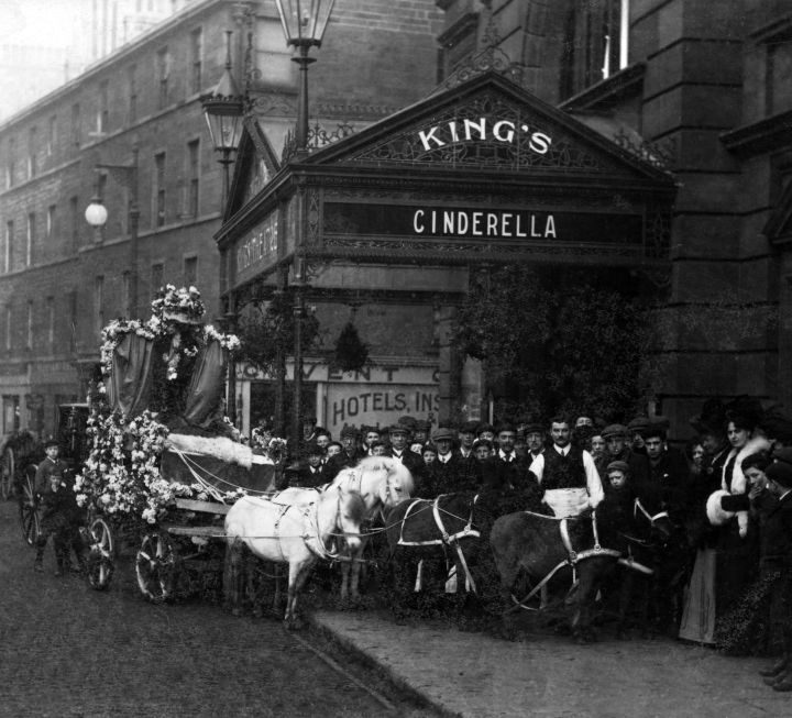 A horse-drawn carriage decorated with flowers on a city street. People are gathered around, and a theatre sign reads "King's Cinderella" above the entrance.