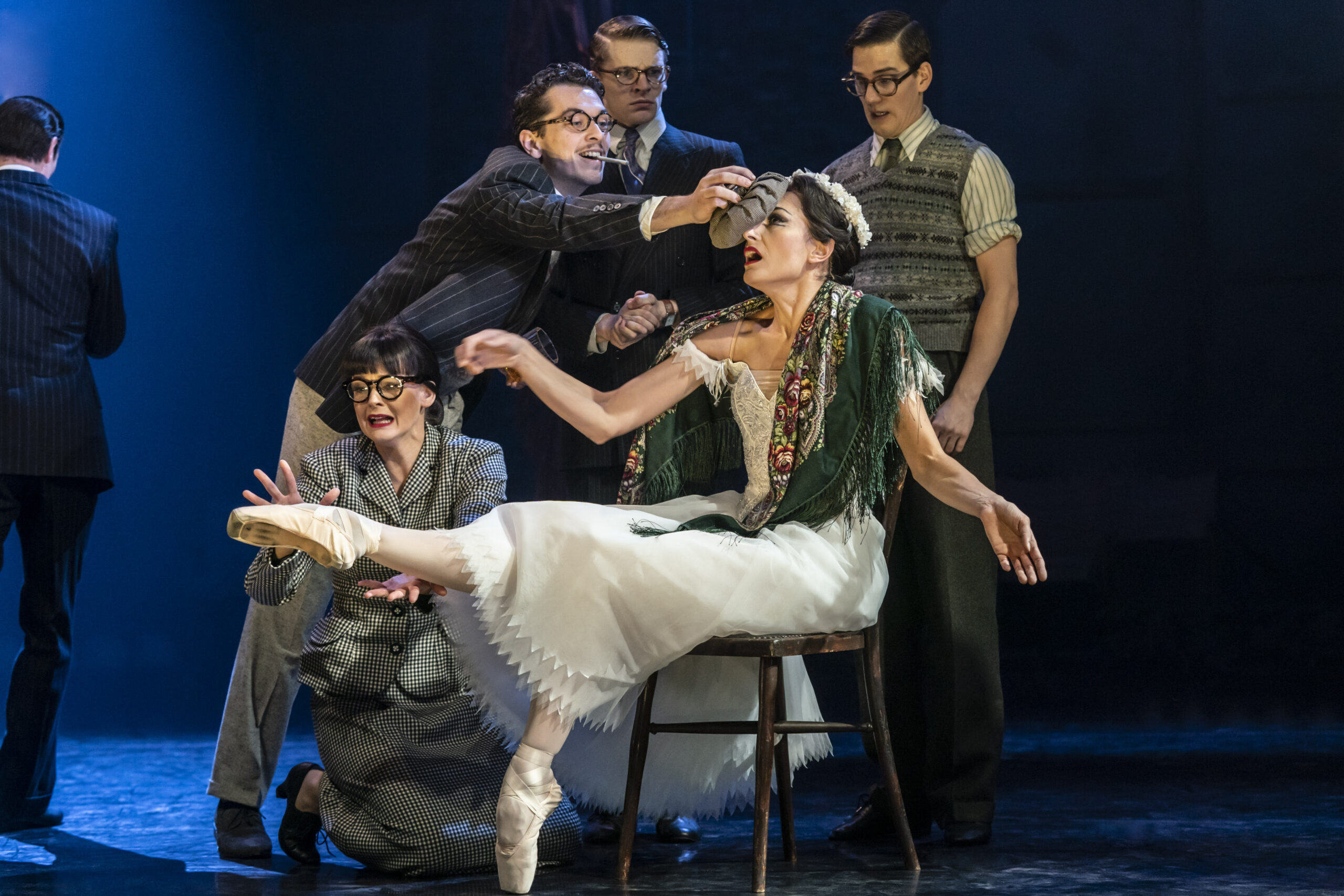 A group of actors in vintage costumes surround a seated ballerina on stage; one man reaches toward her head as she balances en pointe on one foot.