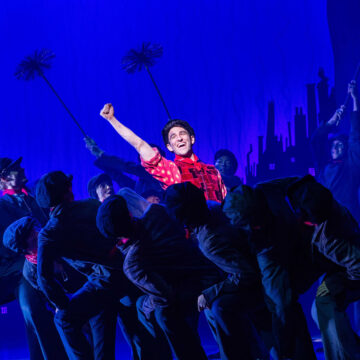 A group of performers dressed as chimney sweeps dance onstage, with one person in a red shirt at the center raising their fist under blue stage lighting.