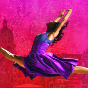 Ballet dancer in a purple dress performs a split leap against a vibrant pink and red cityscape background.