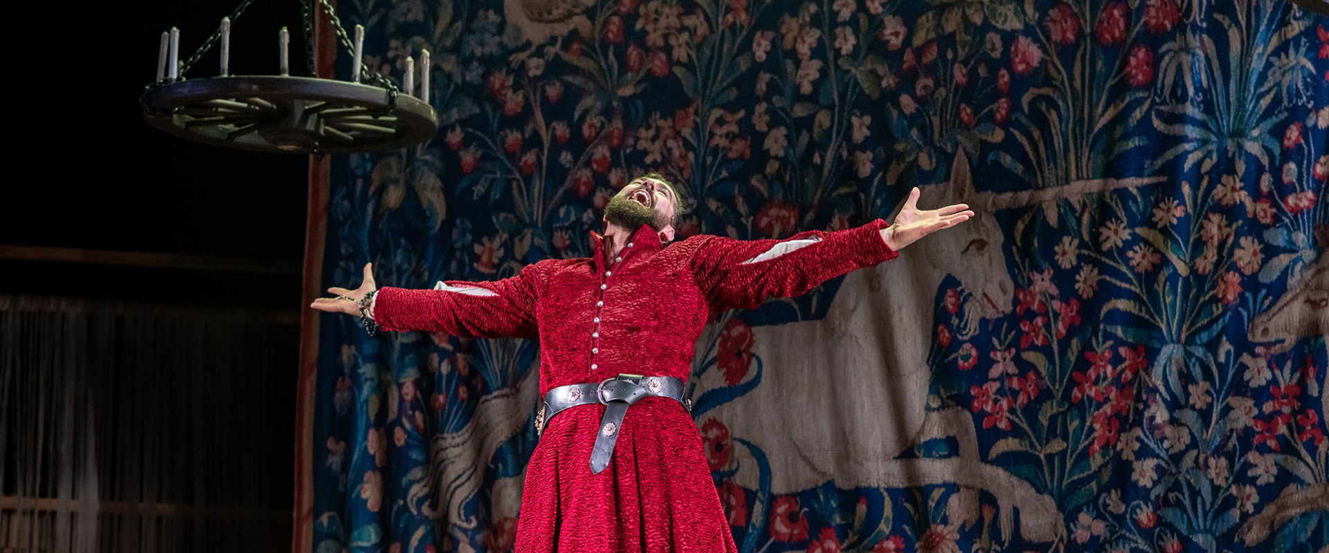 A person in a red medieval costume stands with arms outstretched on stage in front of a tapestry depicting a unicorn.