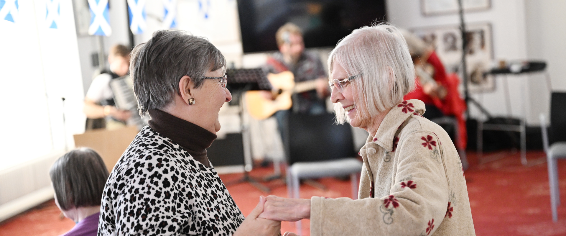 Two elderly women are dancing and smiling indoors. In the background, musicians are playing instruments. Scottish flags are visible in the decor.