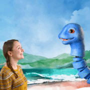 A woman in a yellow sweater smiles at a large blue puppet resembling a sea creature, with an illustrated ocean and mountains in the background.