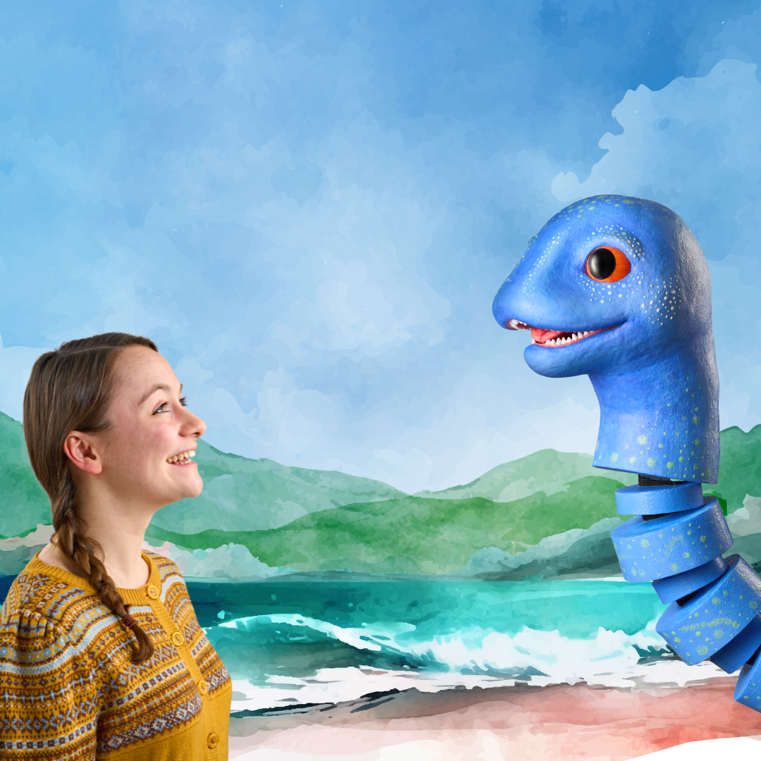A woman in a yellow sweater smiles at a large blue puppet resembling a sea creature, with an illustrated ocean and mountains in the background.