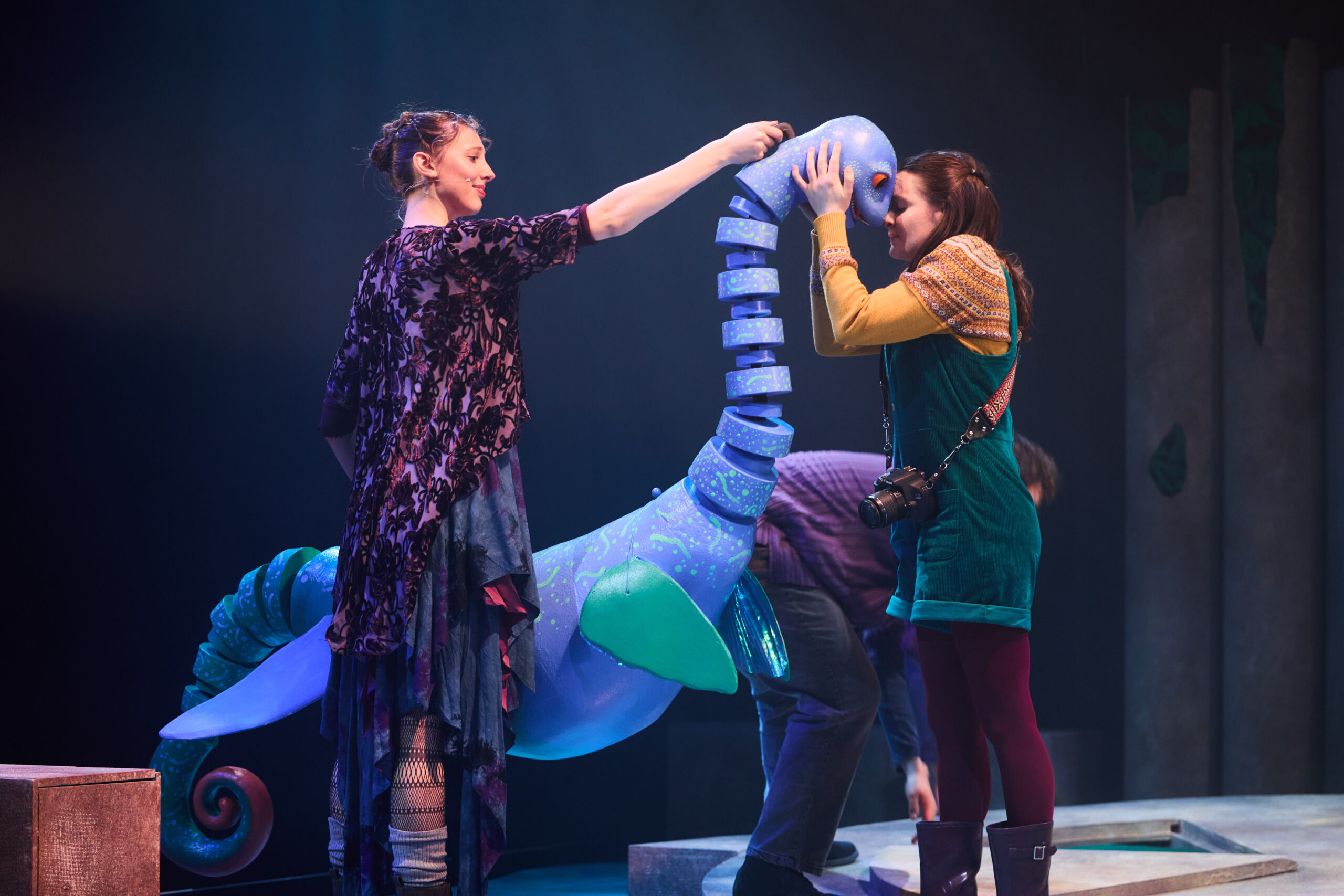 Two women interact with a large blue sea creature puppet on stage during a theatrical performance.