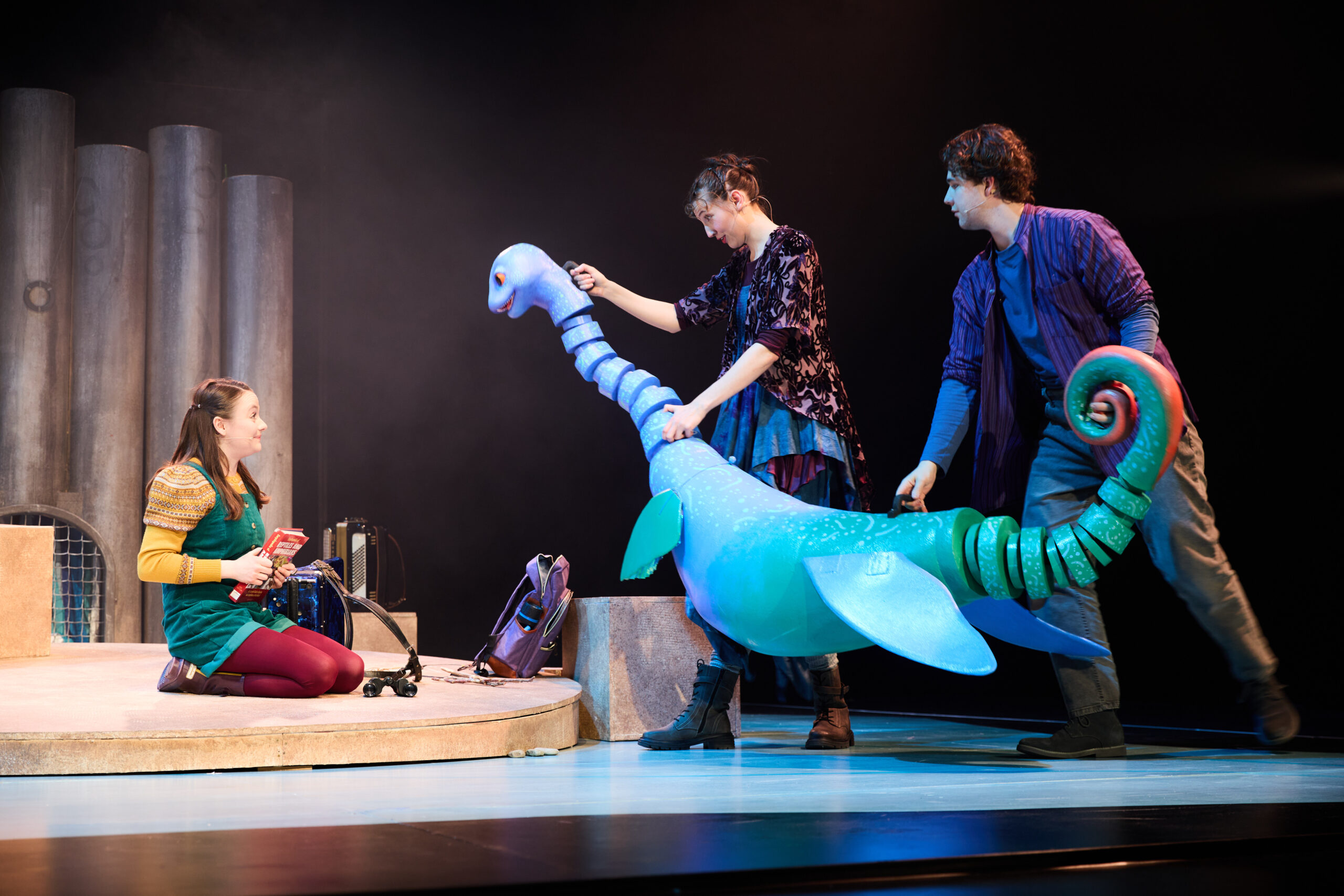 Three actors on stage interact with a large, blue puppet during a theatrical performance with minimal set pieces in the background.