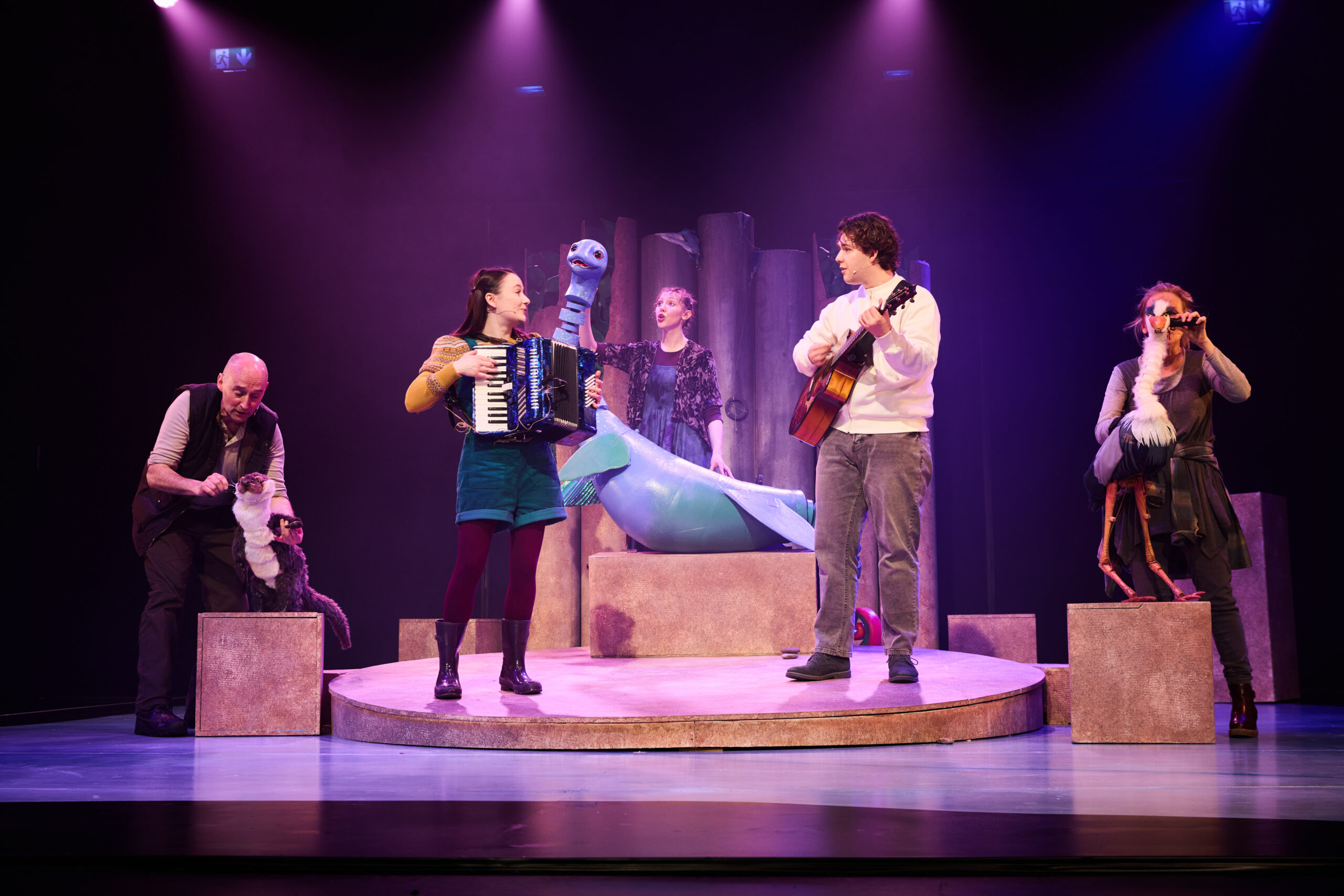 Five performers on stage with puppets; two play musical instruments, one stands behind a large blue puppet, and others operate smaller animal puppets. Stage is lit with purple and pink lighting.